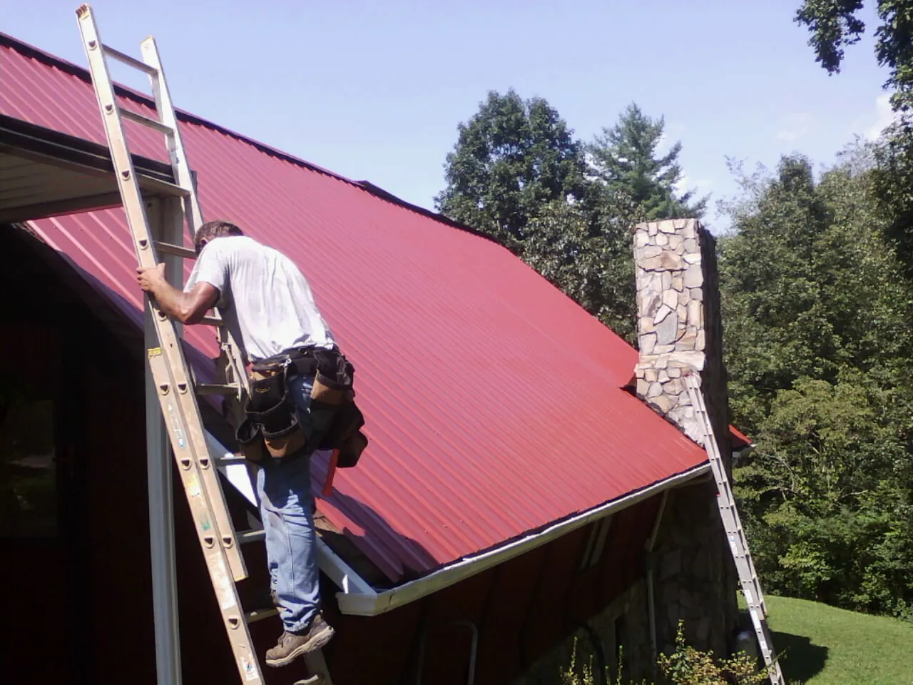 Roofer on a ladder beside a red metal roof and stone chimney — Soffit & Fascia Repair by Roofing Brothers in Pawlet